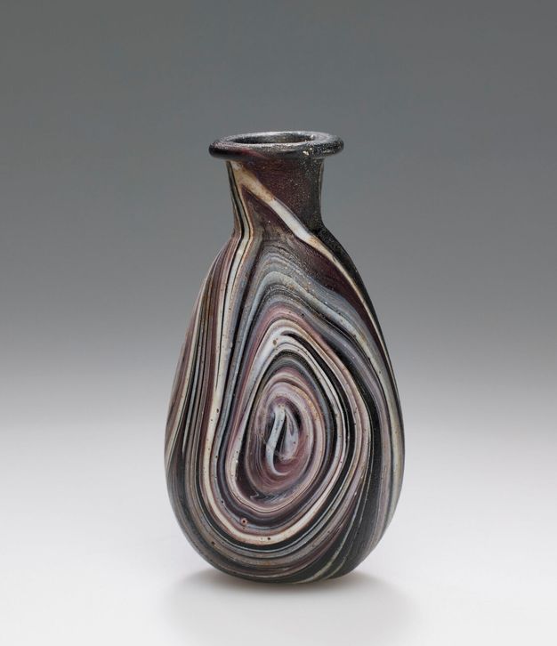 A vase on a plain, gray background. The vase is made of swirls of opaque glass in shades ranging from dark purple or black to lighter purple and white.